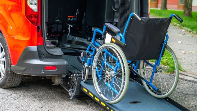  Foto eines leuchtend blauen Rollstuhls, der auf einer Rampe zum Heck eines roten Kleinbusfahrzeuges steht