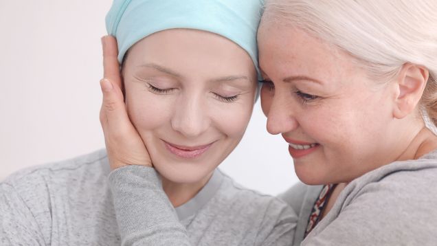 Foto einer leicht lächelnden Frau mit geschlossenen Augen und Krebs-Kopftuch, die von einer anderen Frau mit der Hand sanft Kopf an Kopf gehalten wird
