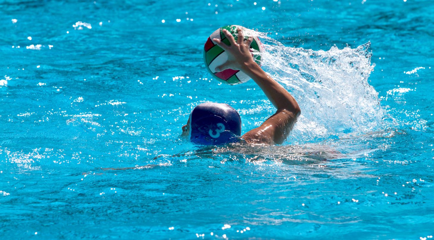 Foto eines Wasserballers im himmelblauen Schwimmbecken, der den Arm mit dem Ball in der Hand zum Wurf aus dem Wasser zieht